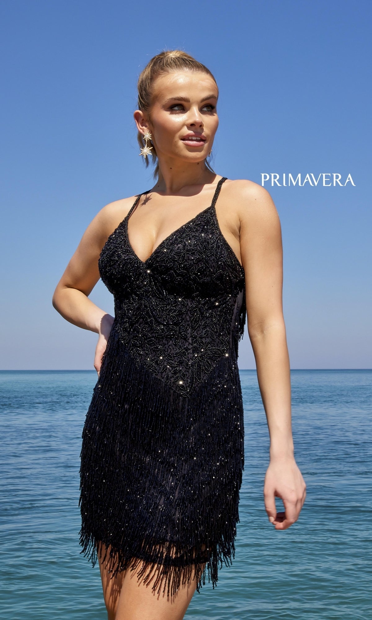 A woman in a Beaded Short Cocktail Dress with Fringe 4045 stands by the ocean under a clear blue sky.